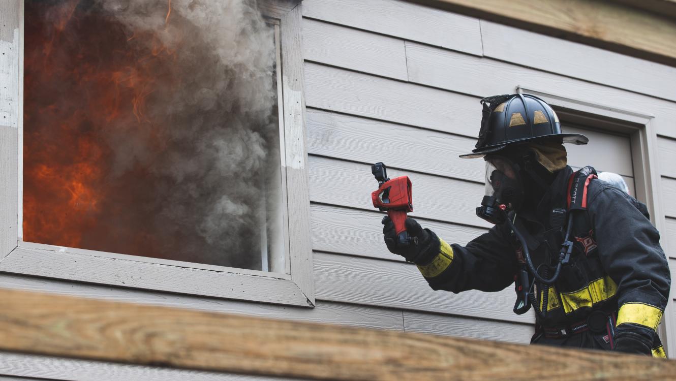 Firefighter holding thermal imaging camera up to a window with smoke coming out