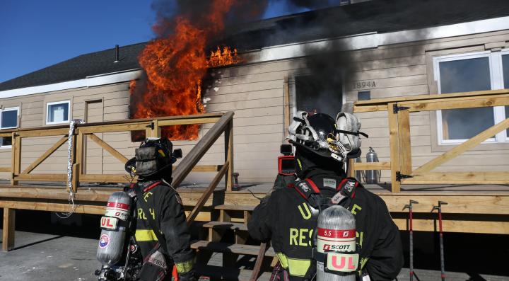 Two firefighters in black firefighter gear standing in front of a house with fire coming out of the left window