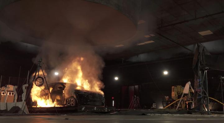 Full-scale vehicle fire safety experiment showing a car engulfed in flames with thick smoke rising. A particulate matter sampler is visible near the burn site, collecting smoke for analysis