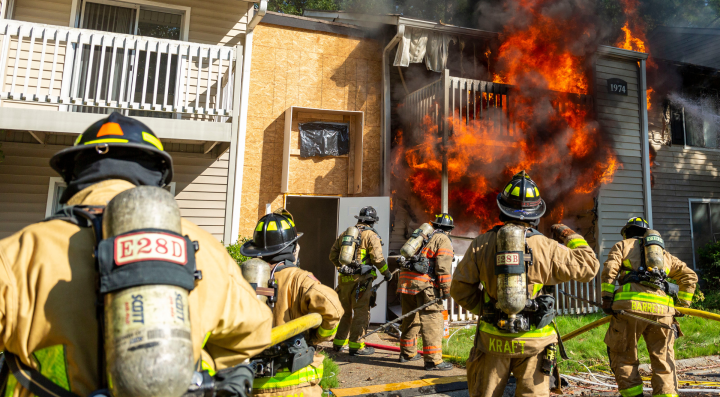 Six firefighters seen from behind assess a fire in a multi-story apartment building while advancing hoselines from the ground. Smoke and flames are visible from multiple units as crews coordinate fire attack operations, reflecting concepts taught in the Evidence-Based Structural Firefighting Course.