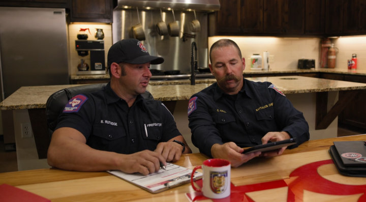 Two firefighters sit at a kitchen table in a fire station, reviewing firefighting training together on a tablet held by one firefighter while both look at the screen.