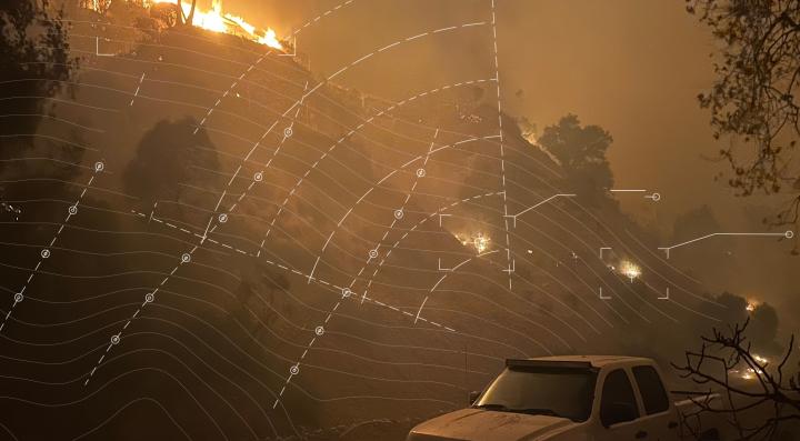 Wildfire spreading across a hillside at night with white contour lines and markers overlaid on top representing wildfire risk modeling