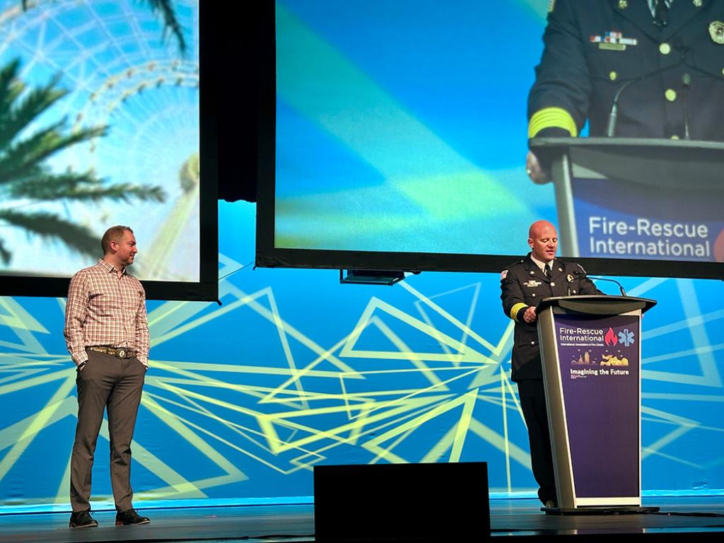 Craig Weinschenk on stage while Josh Waldo speaks at a podium