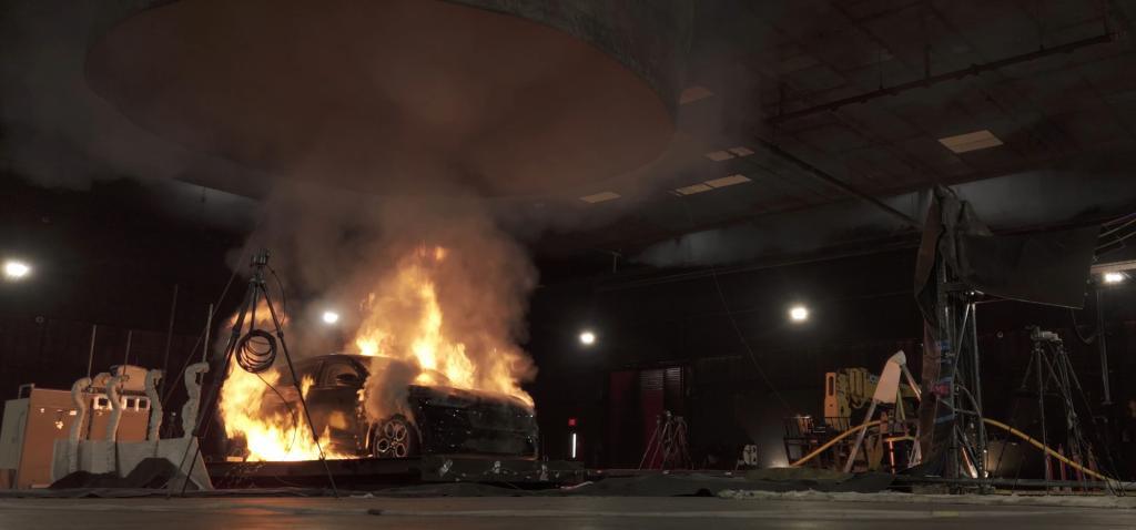 Full-scale vehicle fire safety experiment showing a car engulfed in flames with thick smoke rising. A particulate matter sampler is visible near the burn site, collecting smoke for analysis