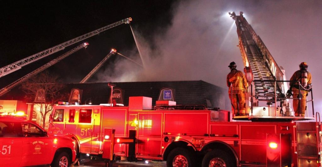 Chula Vista Fire Department crews respond to a warehouse fire. A ladder truck is visible in the foreground, and four extended ladders surround the warehouse, spraying water onto the structure during a near‑miss incident.