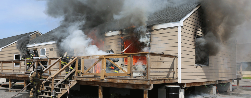 Exterior view of a purpose‑built single‑family test structure during an experimental burn. Side A and part of Side D are visible. A failed window on the right side of Side A is centered in the frame, where a firefighter kneels outside, directing a straight stream into the opening. Smoke billows from every window and door as crews respond.