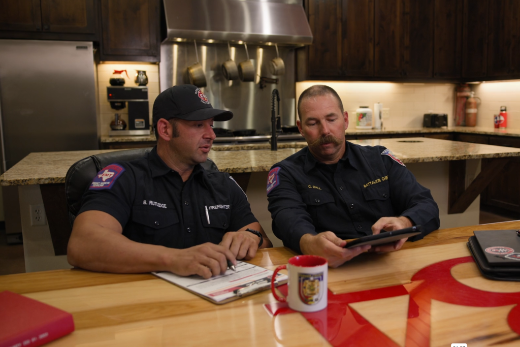 Two firefighters sit at a kitchen table in a fire station, reviewing firefighting training together on a tablet held by one firefighter while both look at the screen.