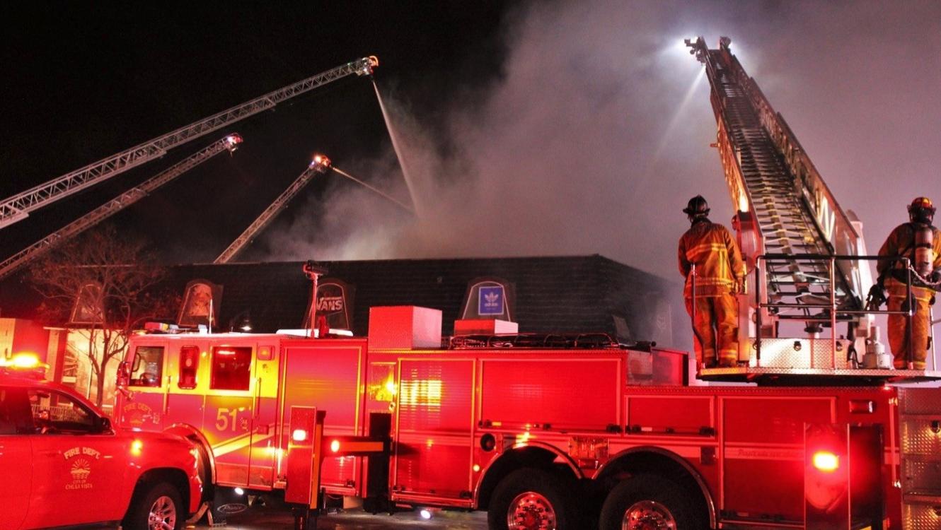 Chula Vista Fire Department crews respond to a warehouse fire. A ladder truck is visible in the foreground, and four extended ladders surround the warehouse, spraying water onto the structure during a near‑miss incident.