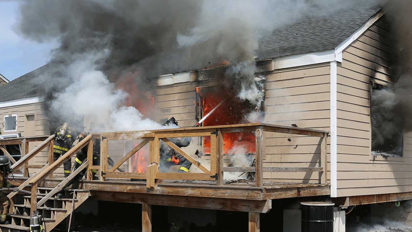 Exterior view of a purpose‑built single‑family test structure during an experimental burn. Side A and part of Side D are visible. A failed window on the right side of Side A is centered in the frame, where a firefighter kneels outside, directing a straight stream into the opening. Smoke billows from every window and door as crews respond.
