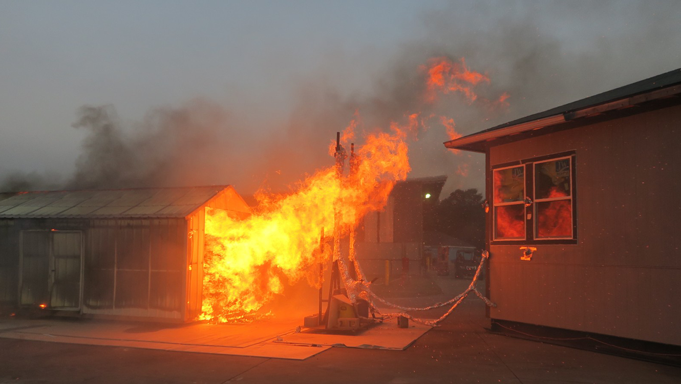 Experiment setup is shown with a test structure, reflective of a California Building Code Chapter 7A-compliant home, exposed to fire from a nearby structure (shed).