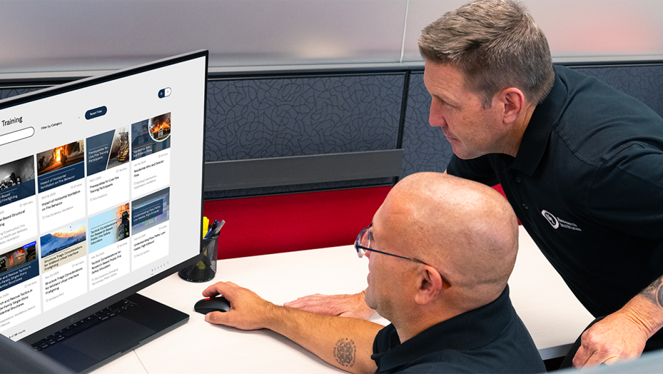 Two firefighters in a cubicle browsing the Fire Safety Academy course library on a computer monitor.