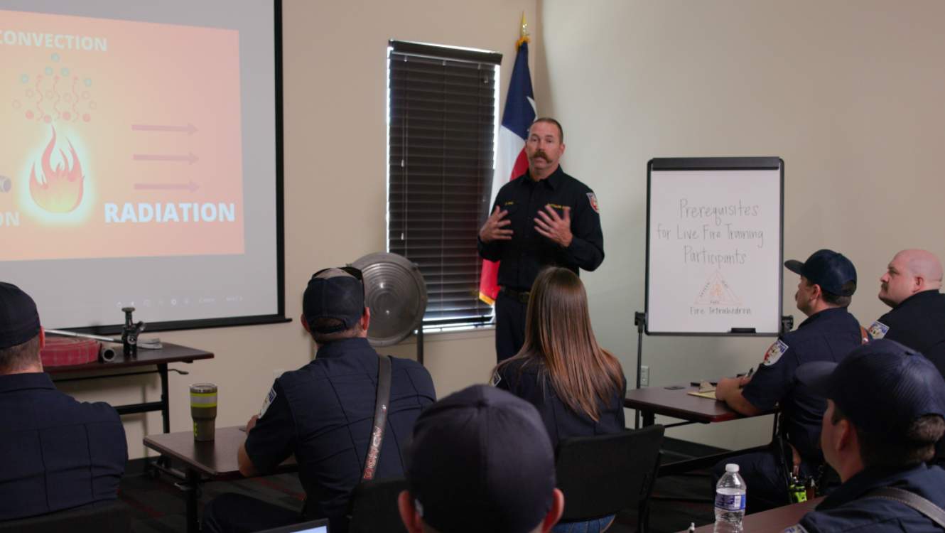 Fire service personnel attend a classroom training session while an instructor presents at the front of the room. Viewed from the back left of the classroom, the image shows the backs of attendees and a presentation screen displaying the fire triangle.