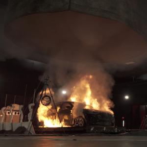 Full-scale vehicle fire safety experiment showing a car engulfed in flames with thick smoke rising. A particulate matter sampler is visible near the burn site, collecting smoke for analysis