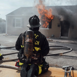 Firefighter in full PPE facing a single‑story home with fire venting from a front window and smoke pushing from the front door.