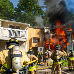 Six firefighters seen from behind assess a fire in a multi-story apartment building while advancing hoselines from the ground.